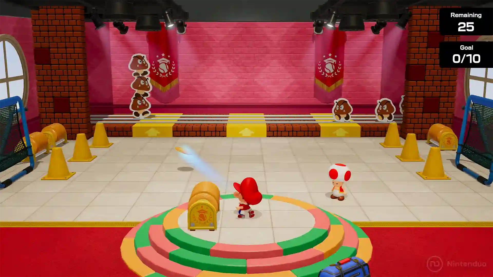 Mario Tennis Fever – mini juego en el que Bebe Mario golpea pelotas de tenis contra barriles y conos amarillos en una pista cubierta, con marcador que indica bolas restantes y objetivo completado.