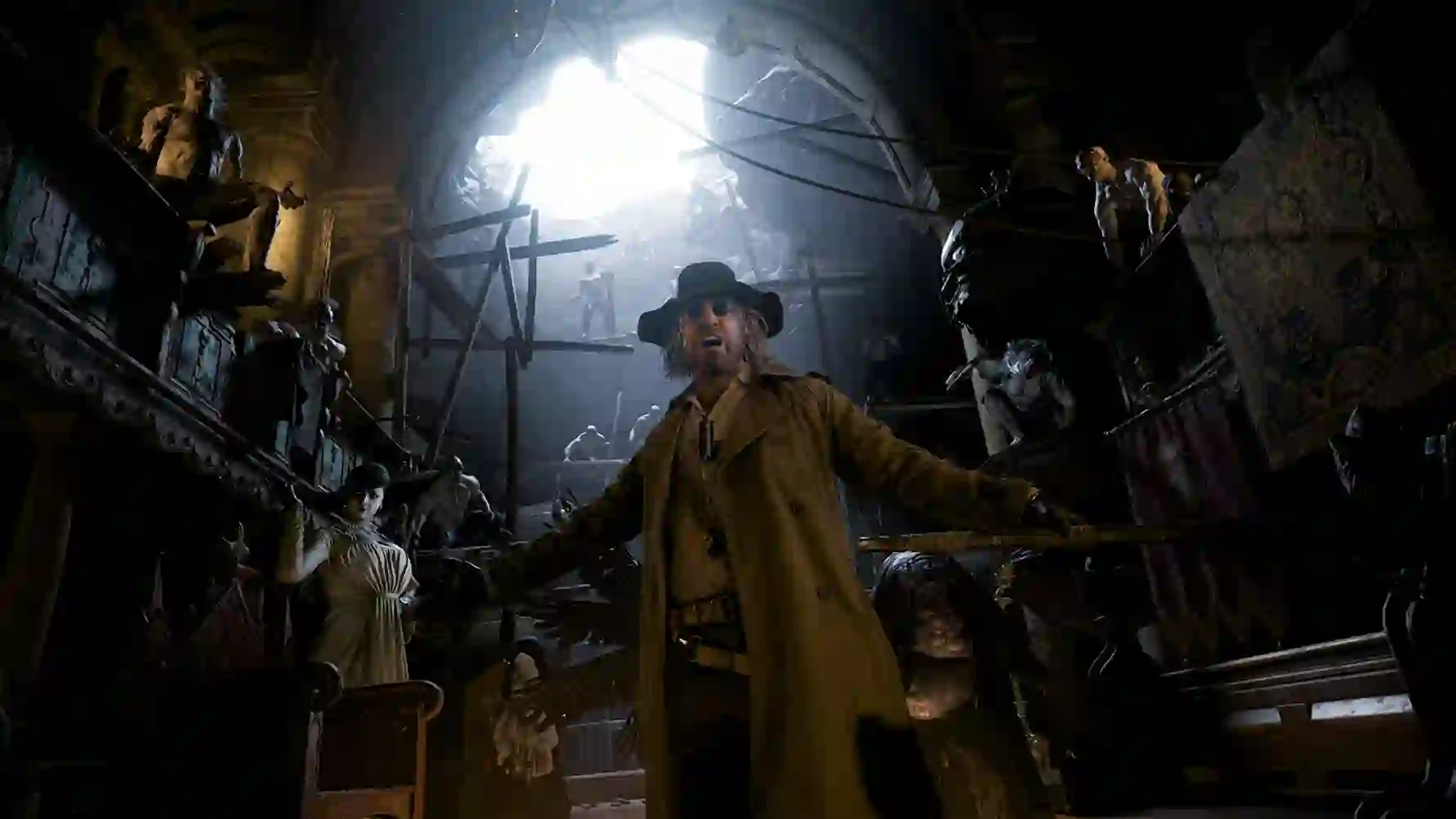 Resident Evil 8 Village: un hombre con sombrero y abrigo largo en una sala oscura llena de estatuas y maniquíes, con luz brillante detrás.