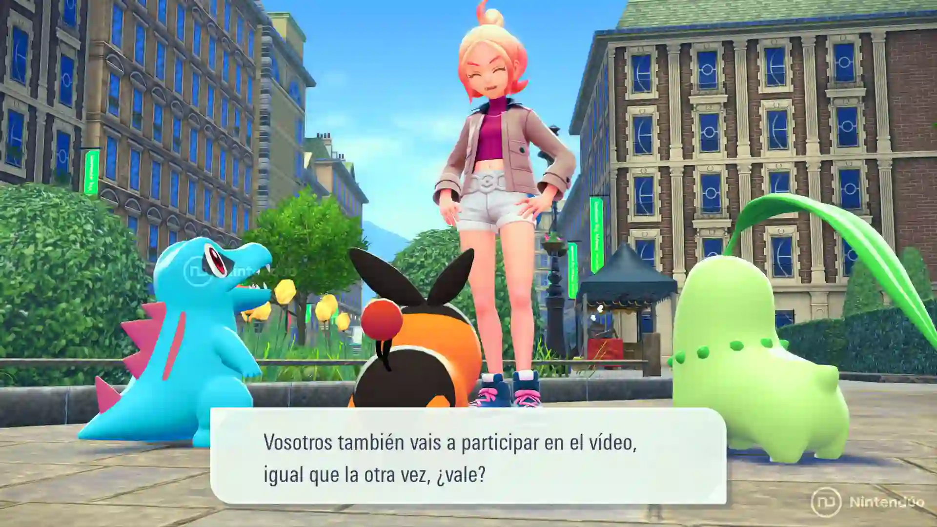 La entrenadora rubia de Leyendas Pokémon Z-A para Nintendo Switch 2 anima a Croconaw, Tepig y Chikorita a participar en un vídeo mientras posan en una plaza de Ciudad Luminalia.
