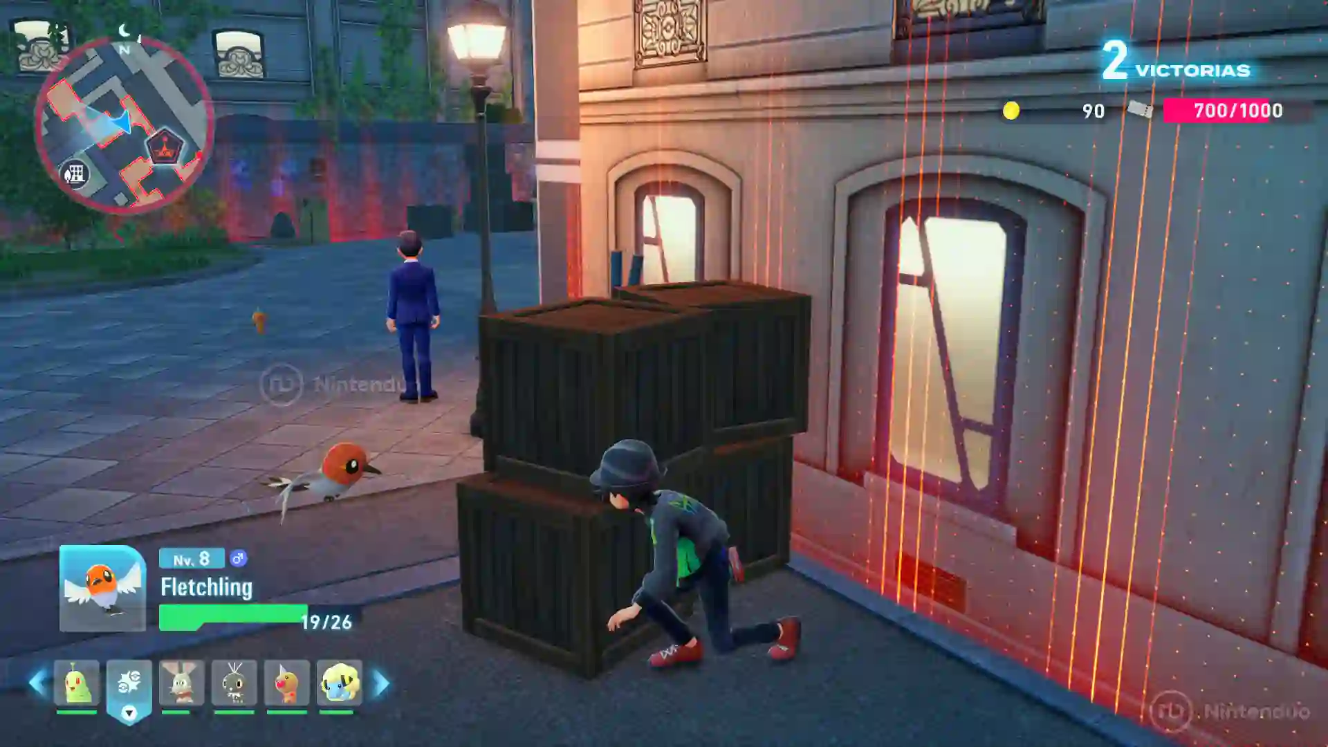 Captura de Leyendas Pokémon Z-A para Nintendo Switch 2 donde el jugador se agacha tras unas cajas de madera para evitar la barrera roja de detección, con un Fletchling a su lado y otro personaje patrullando cerca.